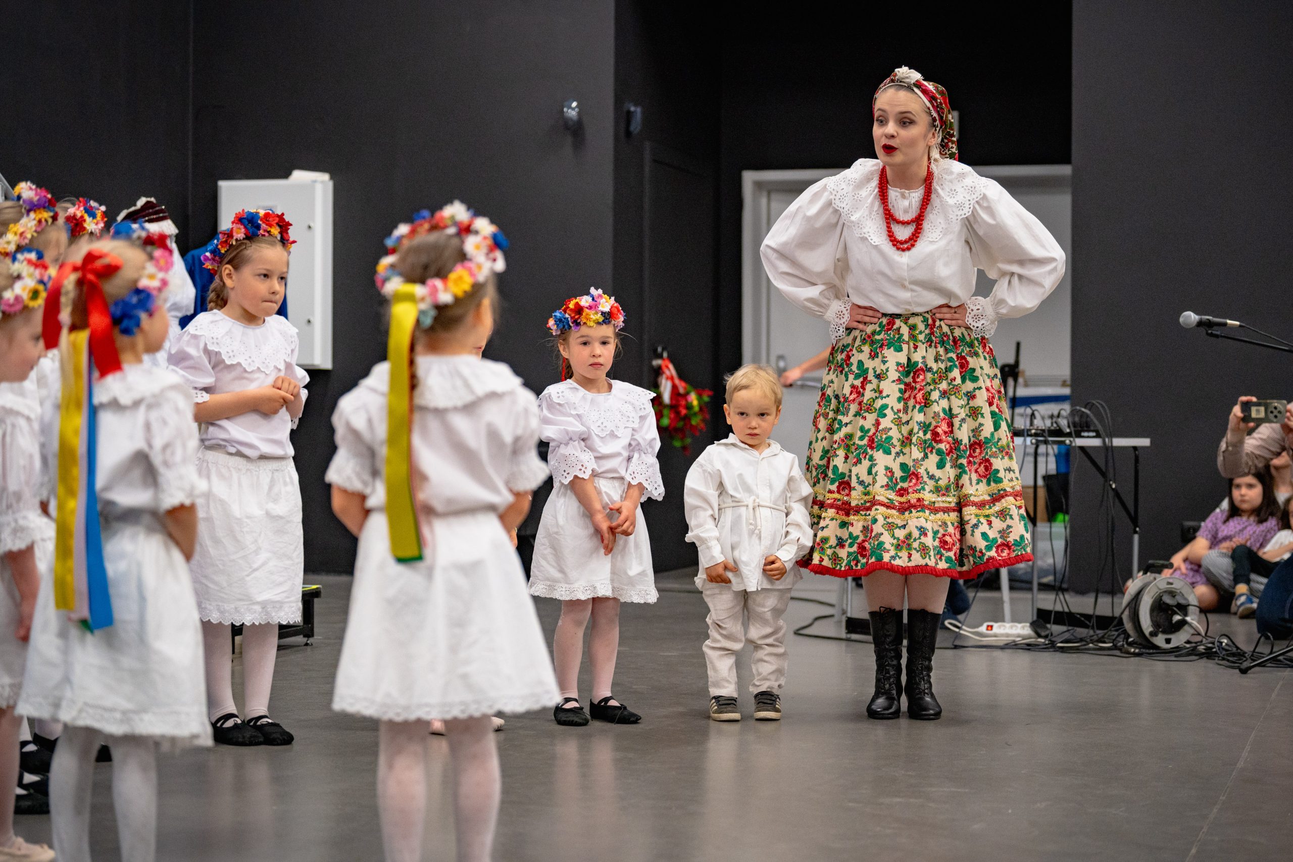dzieci z grupy 3-6 lat w białych strojach stoją w grupie patrząc na instruktorkę i unoszą ręce do góry gestykulując do rymowanki