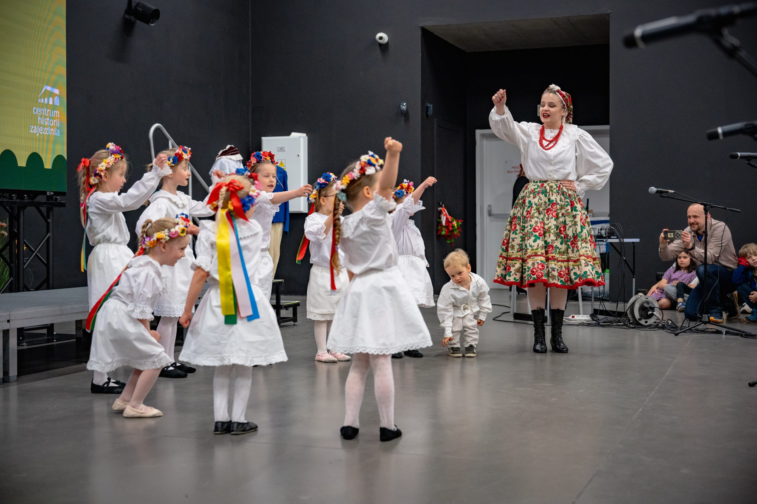 dzieci z grupy 3-6 lat w białych strojach stoją w grupie patrząc na instruktorkę i unoszą ręce do góry gestykulując do rymowanki