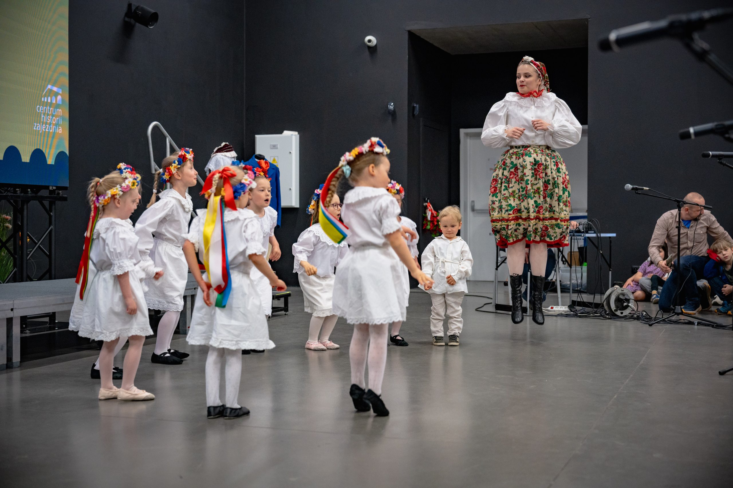 dzieci z grupy 3-6 lat w białych strojach stoją w grupie patrząc na instruktorkę i podskakują do piosenki