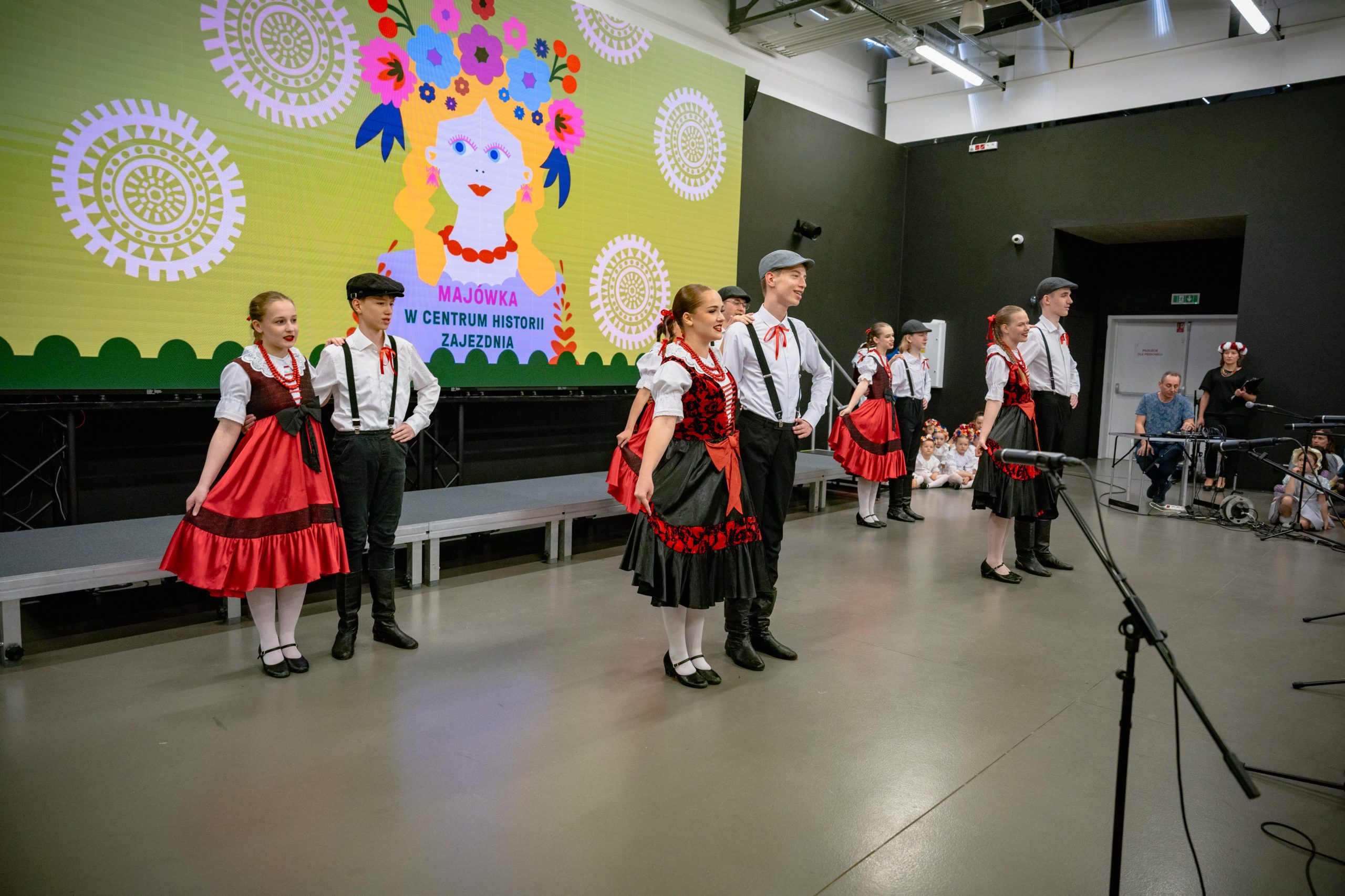 nastolatki w czerwono-czarnych sukienkach oraz chłopcy w białych koszulach, szelkach, czarnych spodniach i kaszkietach w tańcach warszawskich