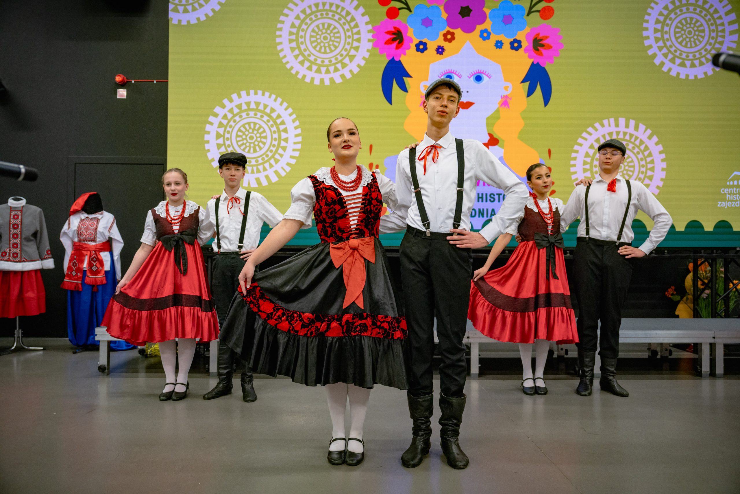 nastolatki w czerwono-czarnych sukienkach oraz chłopcy w białych koszulach, szelkach, czarnych spodniach i kaszkietach w tańcach warszawskich