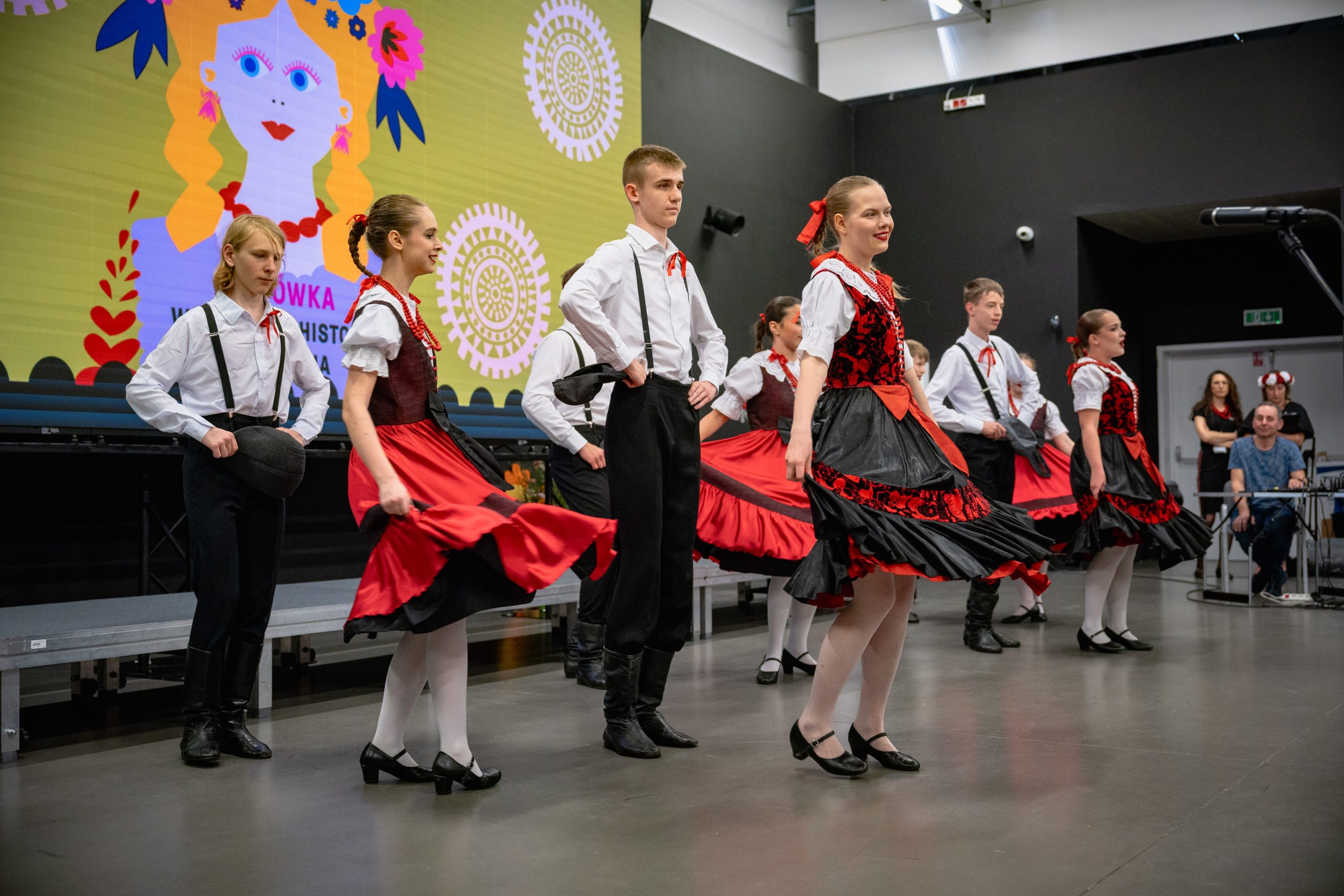 nastolatki w czerwono-czarnych sukienkach oraz chłopcy w białych koszulach, szelkach, czarnych spodniach i kaszkietach w tańcach warszawskich